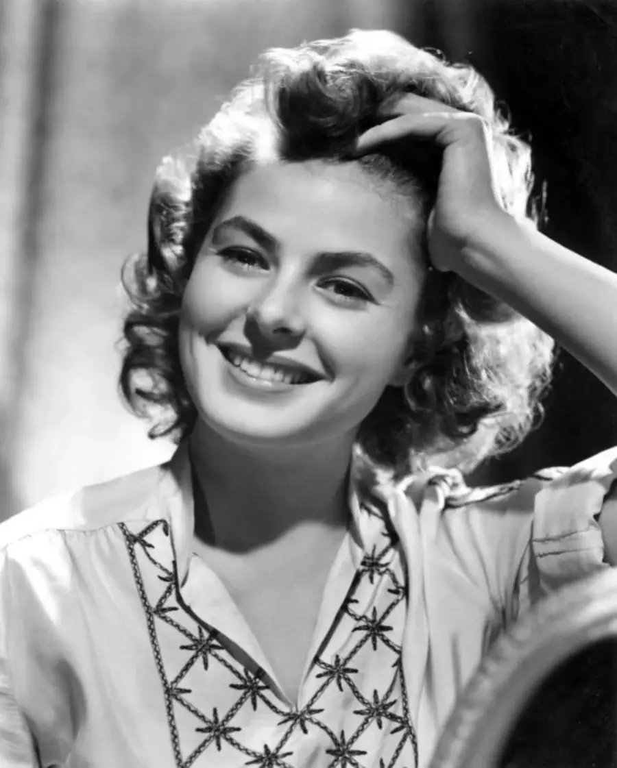 A woman with wavy hair smiles warmly, looking slightly to the side. She is wearing a light blouse with an embroidered pattern and has one hand resting on her head. The photo is in black and white.