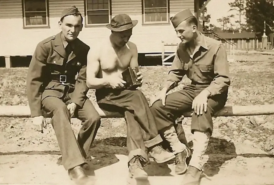 Three young men in vintage military uniforms sit on a wooden rail outside a building; the man in the center is shirtless and examining an object in his hands while the other two men watch.