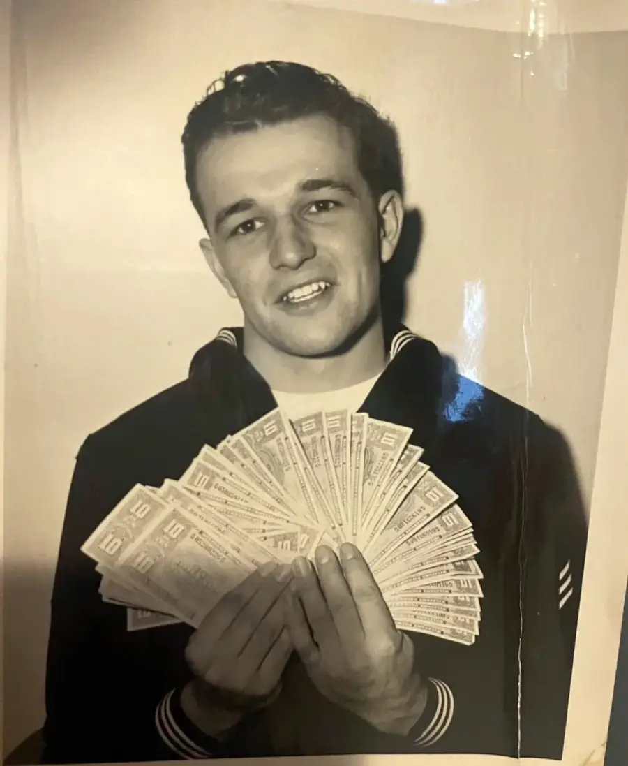 A young man in a dark uniform smiles while holding a large fan of currency bills in both hands, posing in front of a plain backdrop in a black and white photograph.