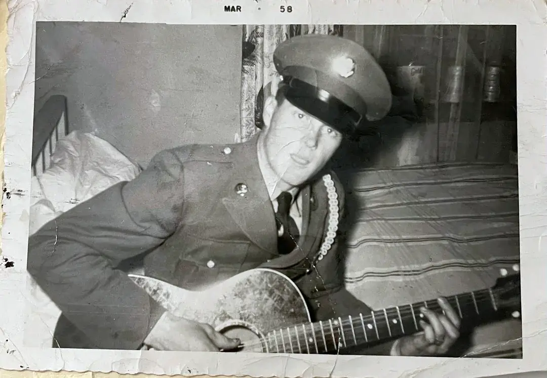 A black-and-white photo from March 1958 shows a man in a military uniform sitting on a bed and playing an acoustic guitar, looking toward the camera. The background includes a striped blanket and a shelf.