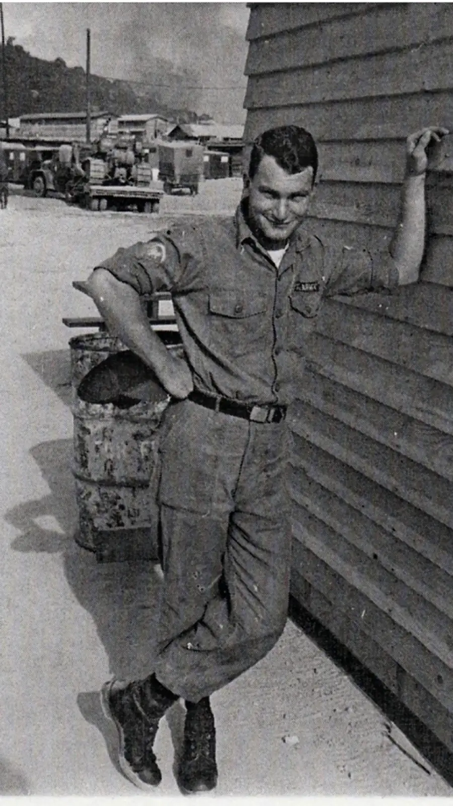 A man in a military uniform leans casually against a wooden building, with one arm resting on the wall. He stands on a dirt path, and there are vehicles and structures visible in the background.