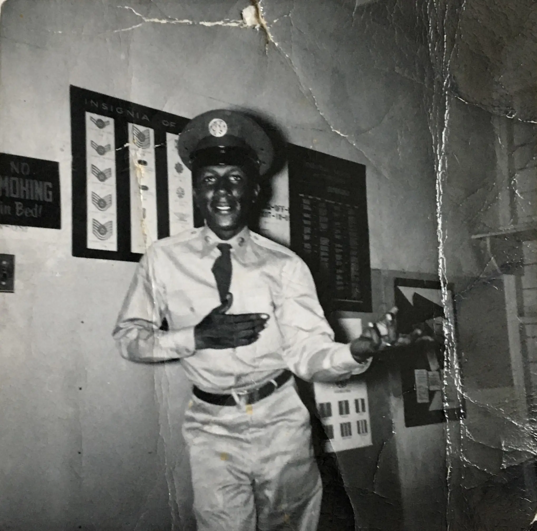 A smiling man in a military uniform and cap stands indoors, gesturing with one hand on his chest and the other extended. Behind him are charts of military insignia and a sign that says “NO MOHING in Bed!”. The photo appears old and worn.