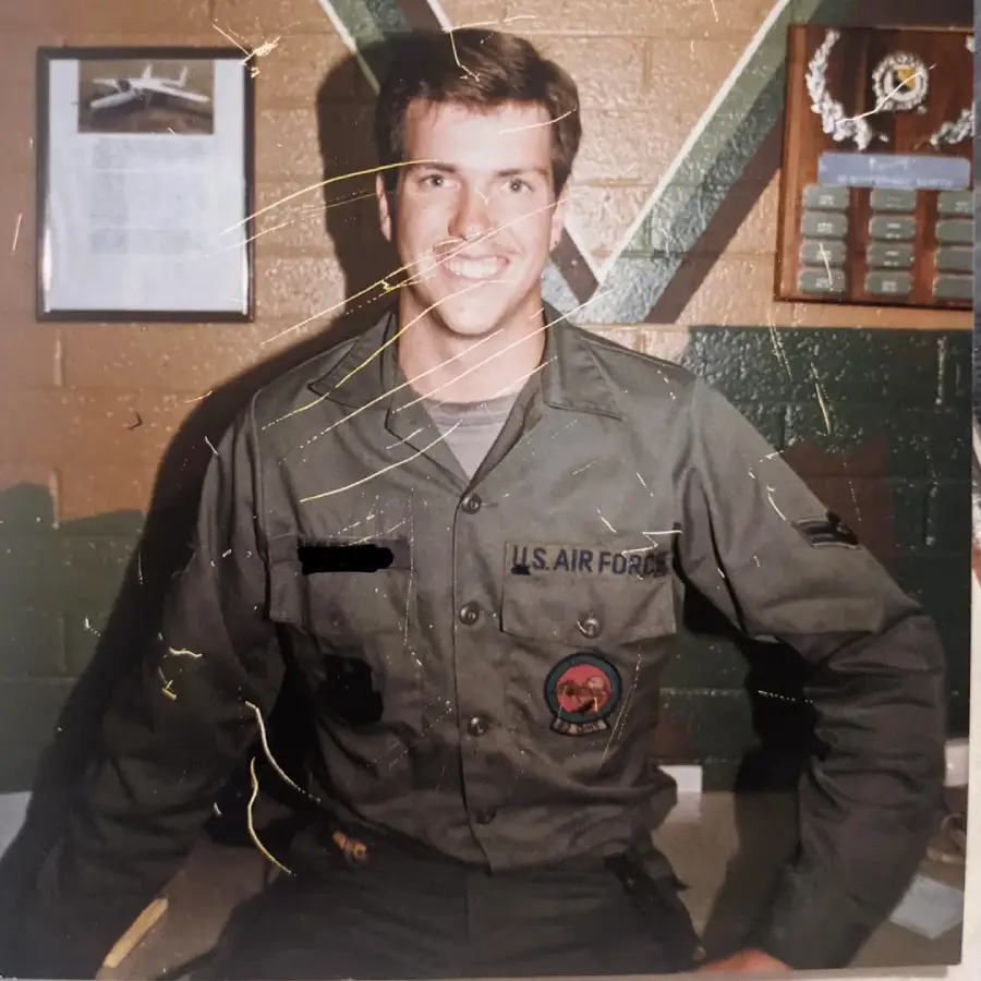 A young man in a U.S. Air Force uniform sits and smiles indoors, with military plaques and a framed photo on the wall behind him. The image shows visible scratches and wear.