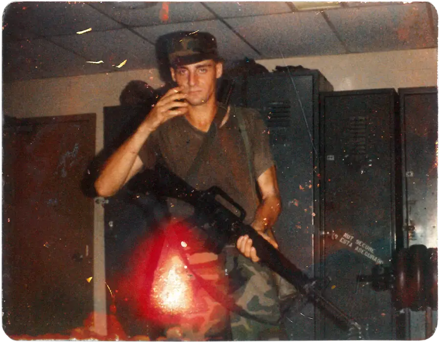 A young man in military uniform holds a rifle and a cigarette, standing in a locker room with dark lockers behind him. The image has visible scratches and signs of aging.