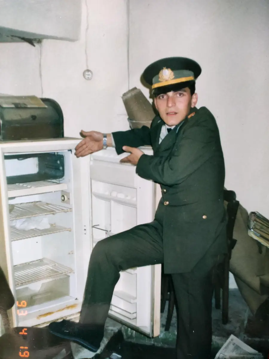 A man in a military uniform gestures dramatically toward an open, nearly empty refrigerator in a cluttered room. The fridge has only a few items inside, and the man appears to be presenting or explaining it.