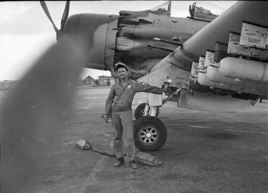 A man in military uniform stands beside the landing gear of a large aircraft on an airfield, with equipment and buildings visible in the background. One of the plane’s propeller blades is blurred in the foreground.