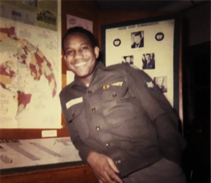 A man in a military uniform smiles and leans against a counter. Behind him are maps and a bulletin board with photos and documents displayed. The scene appears to be indoors, possibly in an office or briefing room.