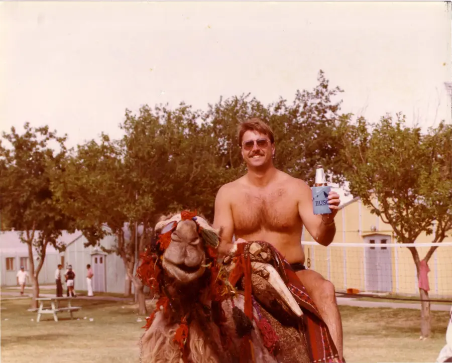 A shirtless man with sunglasses smiles and holds a drink while sitting on a decorated camel in a grassy outdoor area with trees and buildings in the background.