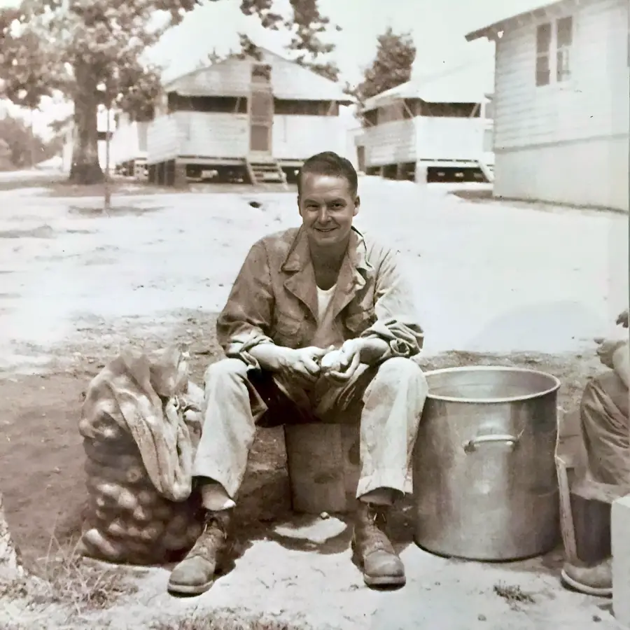 A man in military clothing sits outdoors on a box, smiling while peeling potatoes. A large pot and a bag of potatoes are beside him. Wooden buildings and trees are visible in the background. The photo is in black and white.