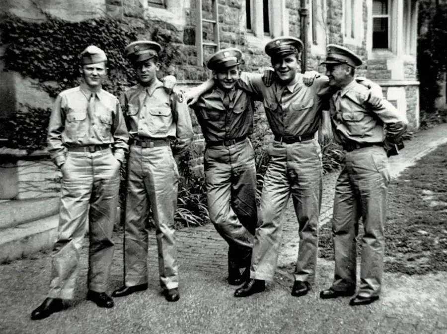 Five men in military uniforms stand arm-in-arm outside a stone building, smiling and posing casually together on a sidewalk lined with greenery. The photo appears to be from the mid-20th century.