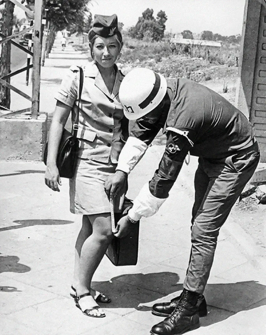 A uniformed officer uses a handheld metal detector to scan a woman’s leg and handbag while she stands on a sidewalk, looking at the camera. The photo is black and white and appears to be from the past.