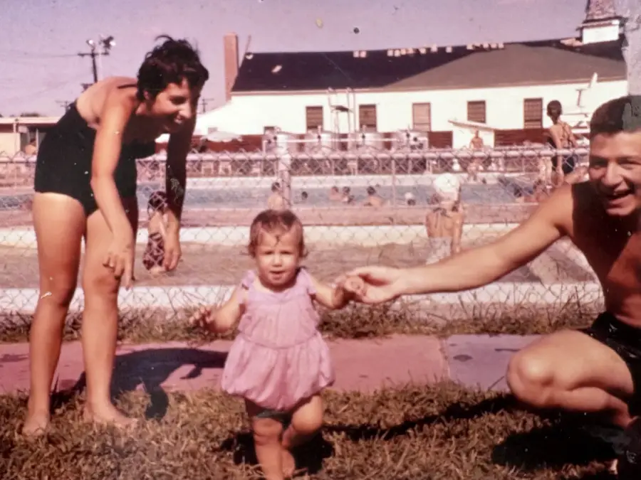 A woman and man in swimsuits help a toddler walk on grass in front of a swimming pool, with people swimming and buildings in the background. The image has a vintage look.