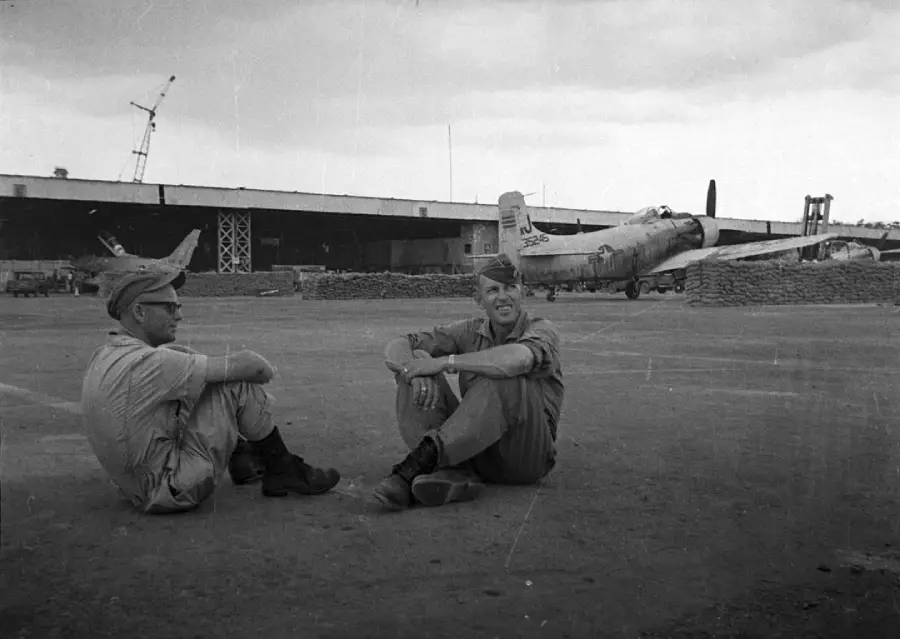 Two soldiers in uniform sit on the ground, talking and smiling, with a military airplane and a large hangar in the background under a cloudy sky.