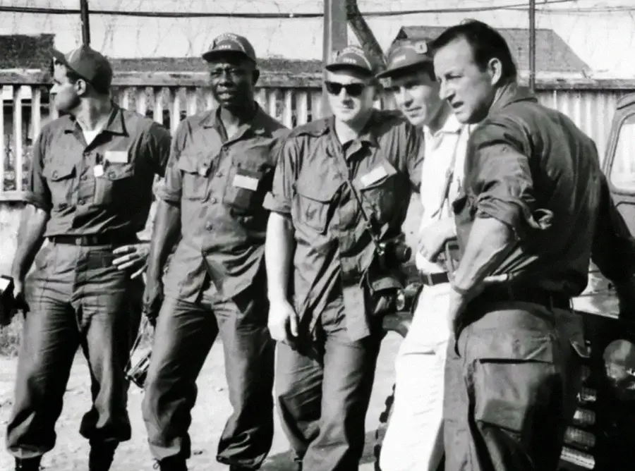 A group of five men in military or paramilitary uniforms stand together outdoors by a fence, some wearing caps and sunglasses, appearing serious and attentive. The image is black and white.