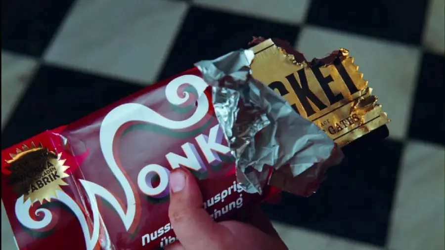 A hand holds a partially unwrapped red "Wonka" chocolate bar, revealing a golden ticket and some chocolate. The background shows black and white checkered tiles.