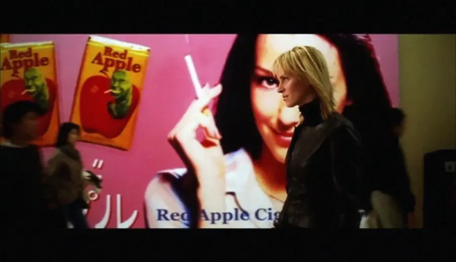 A woman with blonde hair walks past a large, colorful billboard advertising Red Apple Cigarettes, featuring a woman holding a cigarette and two cigarette packs on a pink background. Several blurred people walk nearby.