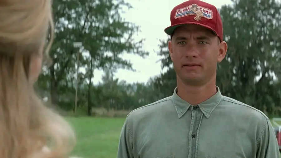 A man wearing a red "Bubba Gump" shrimp cap and a button-up shirt stands outdoors on grass, looking at a blonde woman whose back is to the camera. Trees are visible in the background.