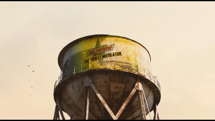 A weathered water tower displays a large yellow and green advertisement for "Brawndo: The Thirst Mutilator" against a hazy sky. The tower is supported by metal legs and a few birds fly nearby.