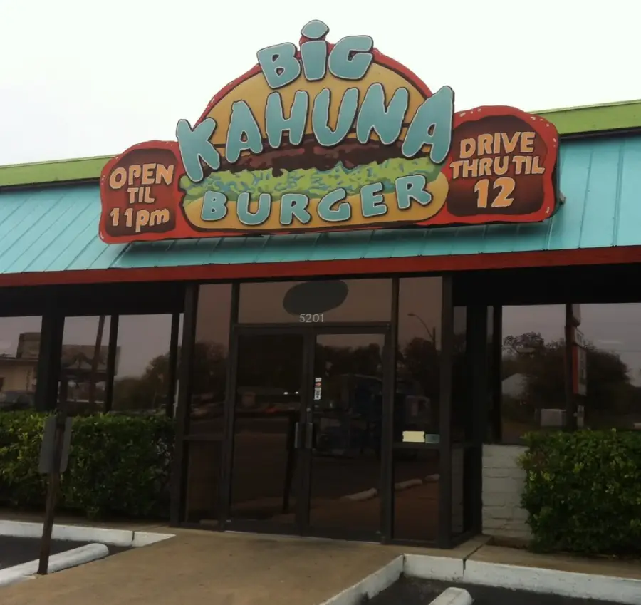 Front view of a Big Kahuna Burger restaurant with a colorful sign above the door, advertising "Open til 11pm" on one side and "Drive Thru til 12" on the other. Glass doors and empty parking spaces are visible.