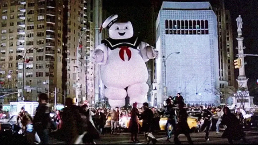 A giant Stay Puft Marshmallow Man in a sailor hat walks through a city street at night as frightened people run away in panic. Tall buildings and streetlights are visible in the background.