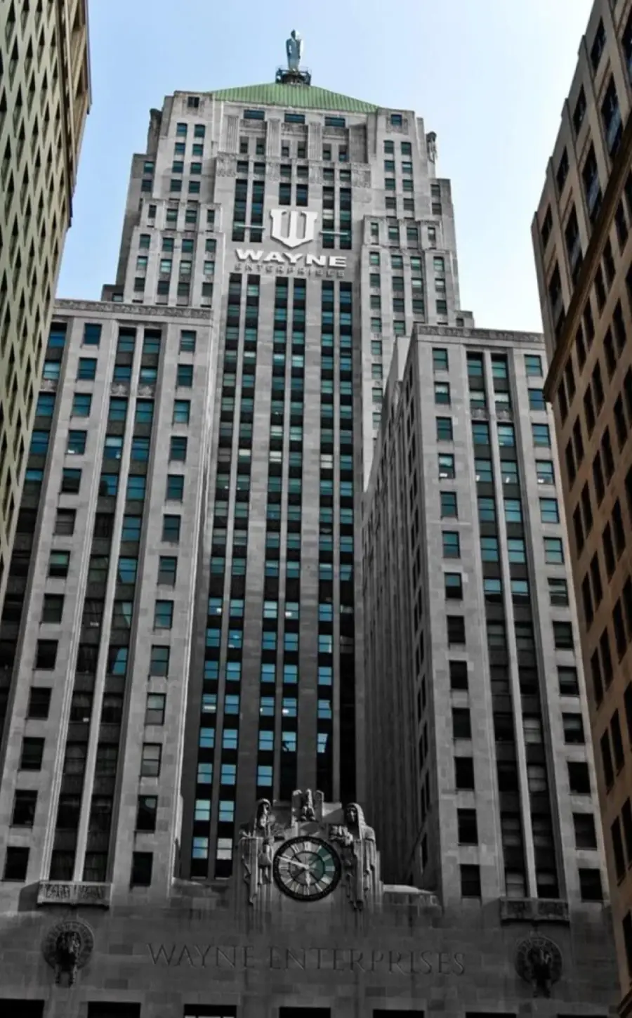 A tall, gray skyscraper with blue-tinted windows and “Wayne” written near the top, flanked by other high-rise buildings, seen from street level against a clear sky.
