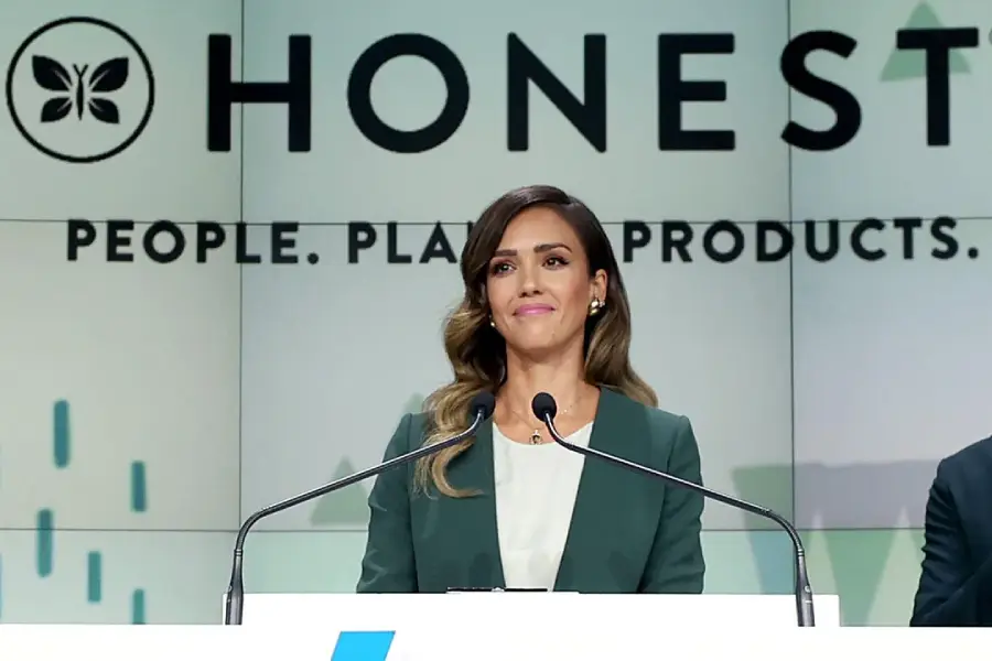 A woman in a green blazer stands at a podium, speaking. Behind her is a large sign that reads "HONEST" and "People. Planet. Products." She appears confident and professional.