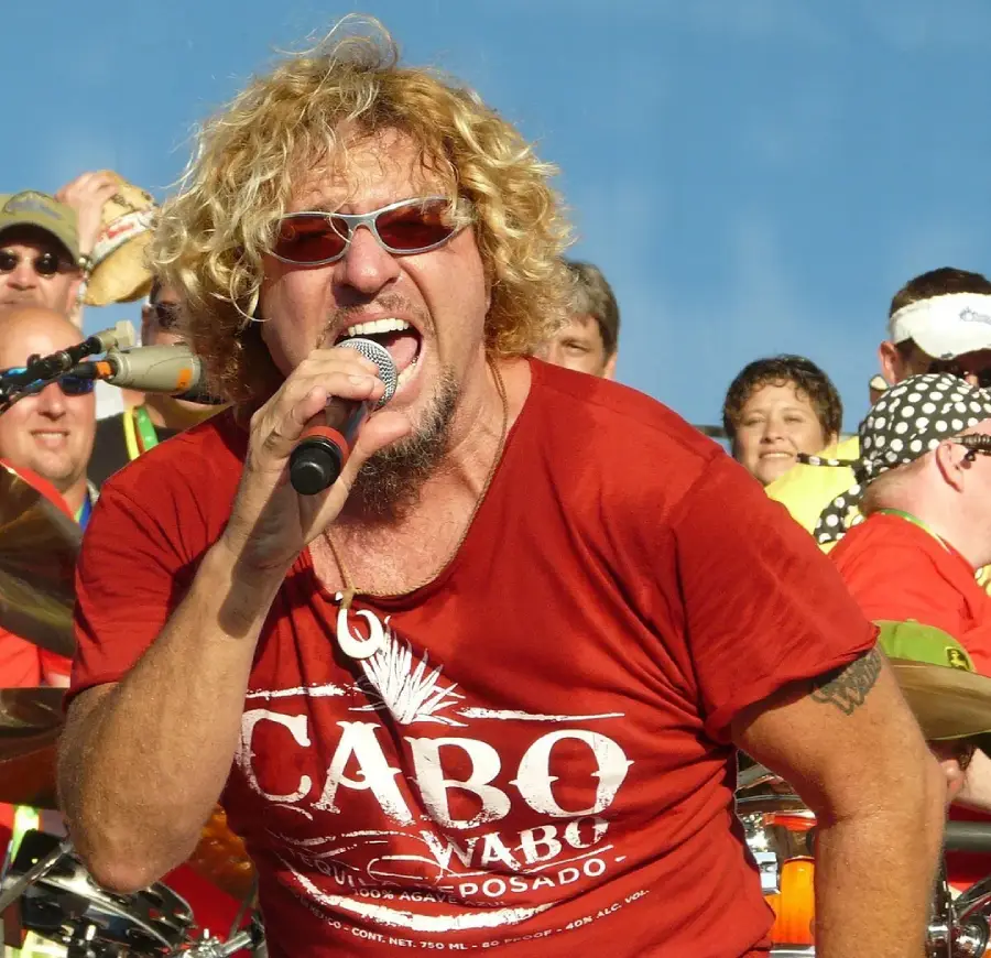 A man with curly blond hair, sunglasses, and a red "Cabo Wabo" shirt passionately sings into a microphone at an outdoor event, with a crowd of people and a drummer in the background.