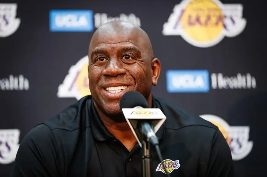 A man wearing a black Lakers polo smiles while speaking into a microphone at a press conference, with a backdrop featuring the Lakers and UCLA Health logos.