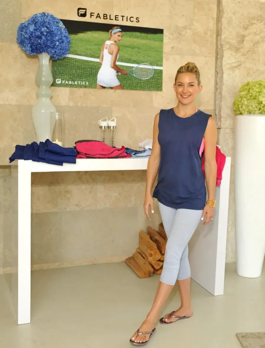 A woman in a blue sleeveless top, light gray leggings, and flip-flops stands by a white table with folded clothes. Behind her is a Fabletics tennis-themed poster and large vases with blue and green flowers.
