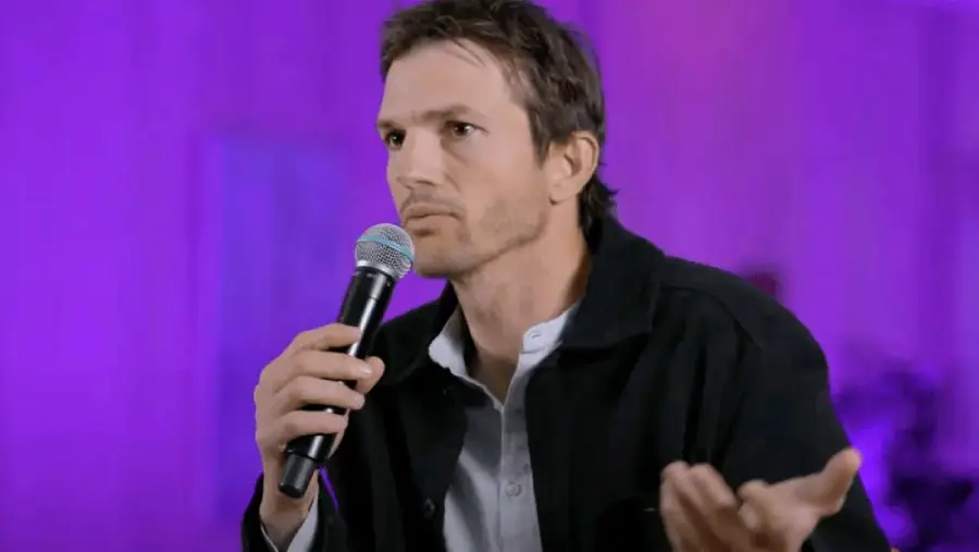 A man with short brown hair holds a microphone and gestures with his other hand while speaking. He is wearing a dark jacket over a light shirt, and the background is lit in purple.