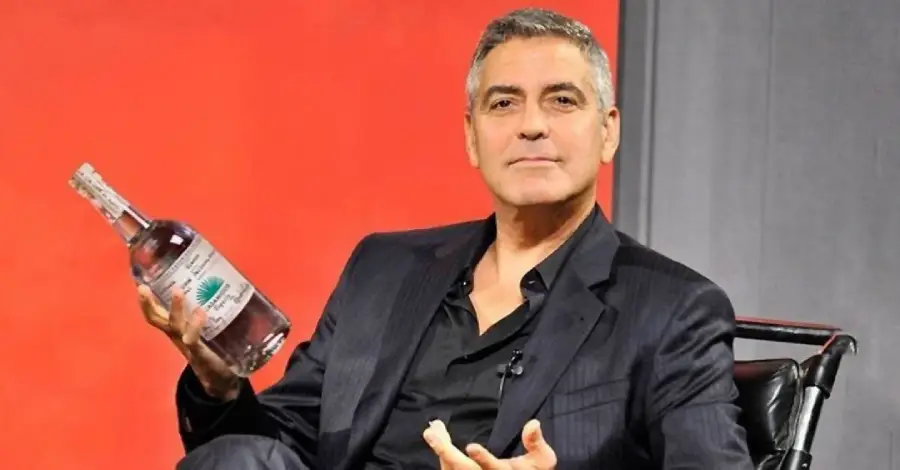A man in a dark suit sits in a chair, holding a clear bottle with a label in one hand and gesturing with the other, against a red and gray background.