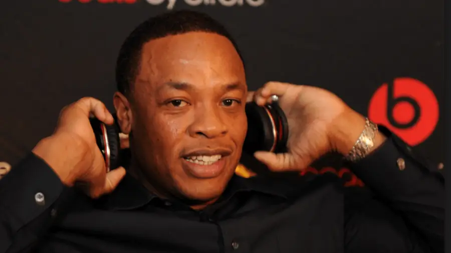 A man wearing a dark shirt holds a pair of headphones to his ears and smiles. The background features the Beats by Dre logo and other branding.