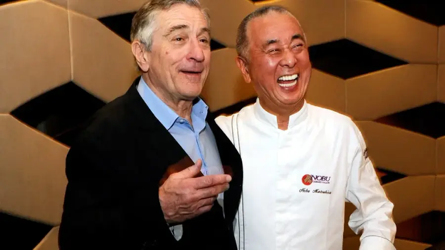 Two older men smile and pose together. The man on the left wears a dark jacket and blue shirt, and the man on the right wears a white chef’s coat with “Nobu” embroidered on it. They stand in front of a geometric-patterned wall.
