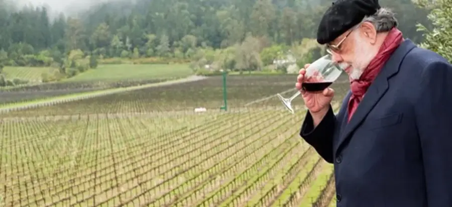 An older man in a beret and scarf holds a glass of red wine, smelling it thoughtfully, while standing next to a large vineyard with rows of grapevines and green hills in the background.