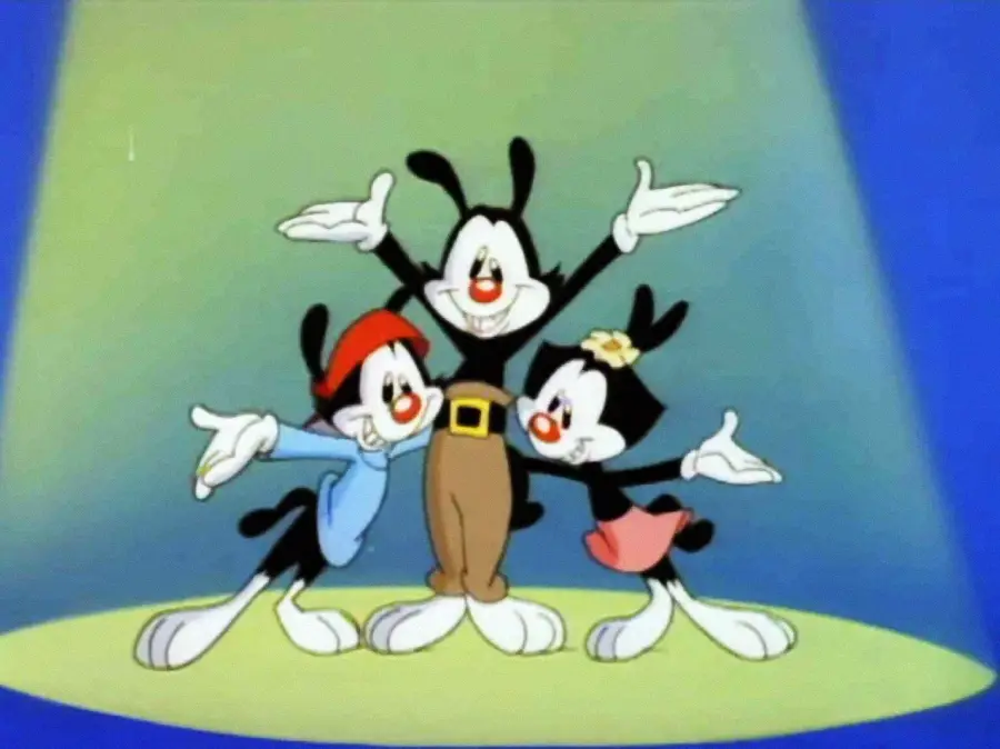 Three cartoon characters with black fur, large white faces, and big ears stand together smiling under a spotlight. One wears brown pants, one a blue shirt and red cap, and the third a pink skirt with a yellow flower on her head.