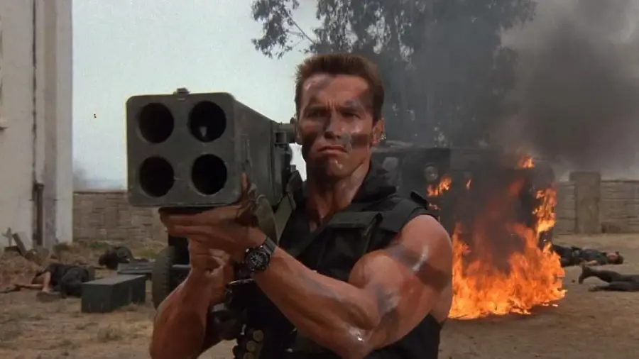 A muscular man with camouflage paint holds a large four-barreled rocket launcher, aiming forward, with fire and smoke rising from a burning vehicle in the background.