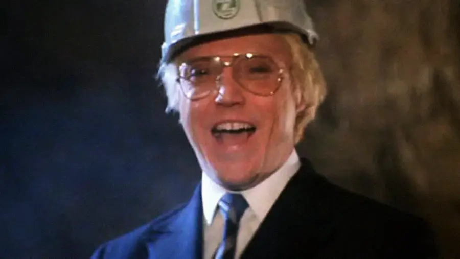 A man wearing a silver hard hat, glasses, and a suit smiles widely. The background is dark and blurred, highlighting his face and attire.