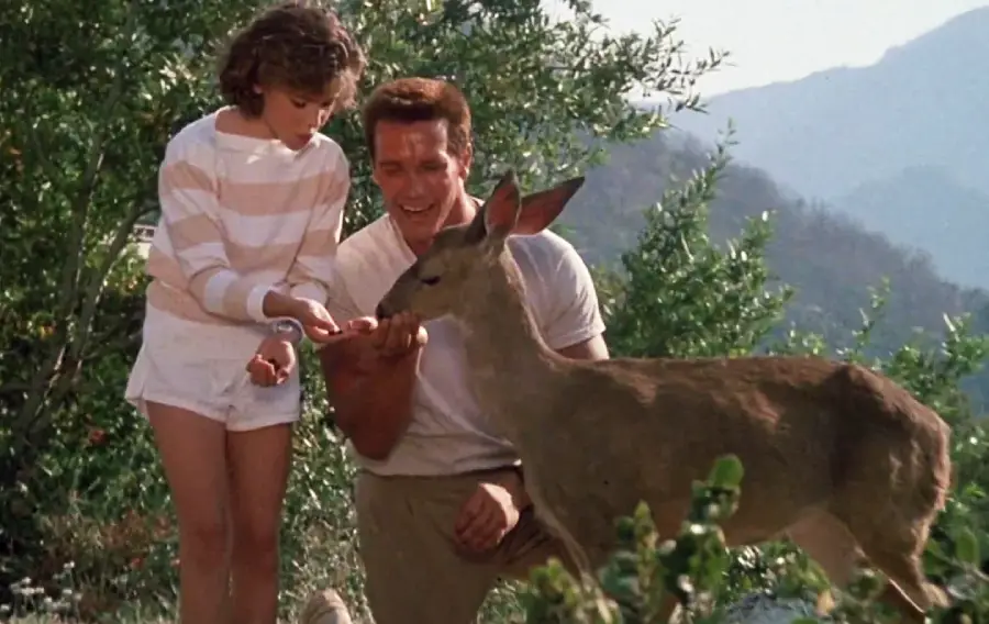 A man and a young girl feed a deer outdoors, surrounded by greenery and mountains under a clear sky. The girl wears a striped shirt and shorts, while the man kneels beside her, smiling.