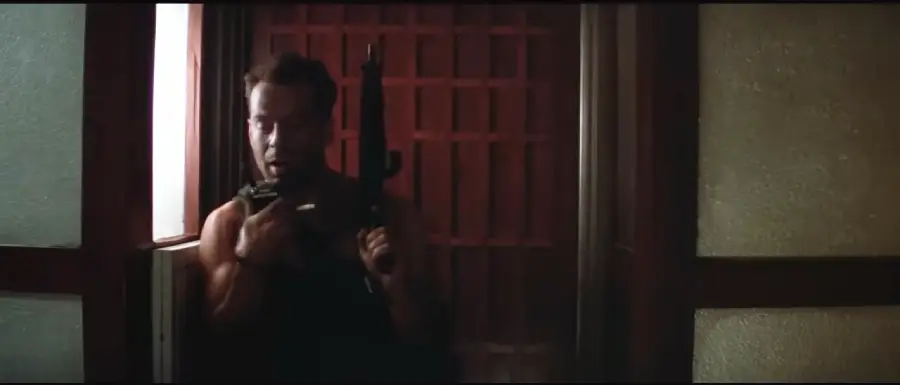 A man in a dark tank top stands by a window, holding a walkie-talkie in one hand and a rifle in the other, with a tense expression on his face. The background is dimly lit with red and frosted glass panels.