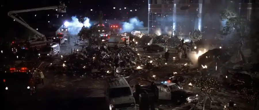 A nighttime scene shows emergency vehicles and firefighters responding to a large debris-strewn area with smoke and small fires, indicating recent destruction or disaster in an urban setting.