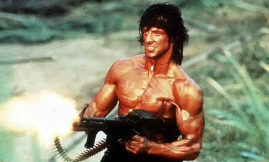 A muscular man with a headband is shirtless, holding and firing a large machine gun in a jungle setting, with an intense expression on his face.