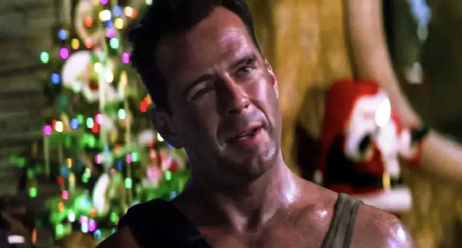A man with short hair and a sweaty face stands indoors in front of a decorated Christmas tree and a Santa decoration, wearing a sleeveless shirt and carrying a shoulder strap.