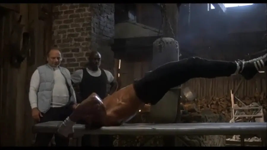 A shirtless man does intense ab exercises on a bench in a rustic gym, while two men stand behind him watching. There are brick walls, wooden beams, and a punching bag in the background.