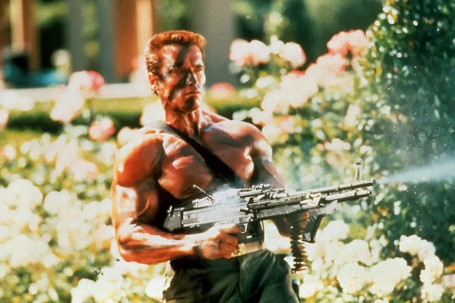 A muscular man with a bare torso and ammo belt fires a large machine gun outdoors, surrounded by greenery and flowers, with an intense expression on his face.
