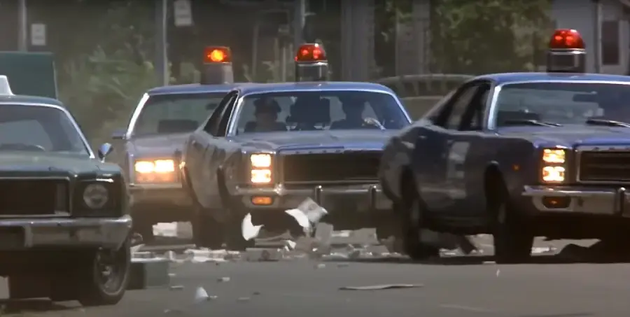 Three blue police cars with red lights flashing speed down a debris-filled street, with paper and objects scattered on the ground, suggesting a high-speed chase or emergency situation.