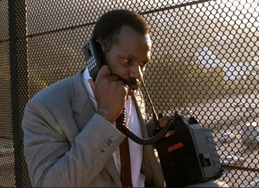 A man in a gray suit stands outdoors by a chain-link fence, holding a large vintage mobile phone and talking with a serious expression. Sunlight filters through the fence in the background.