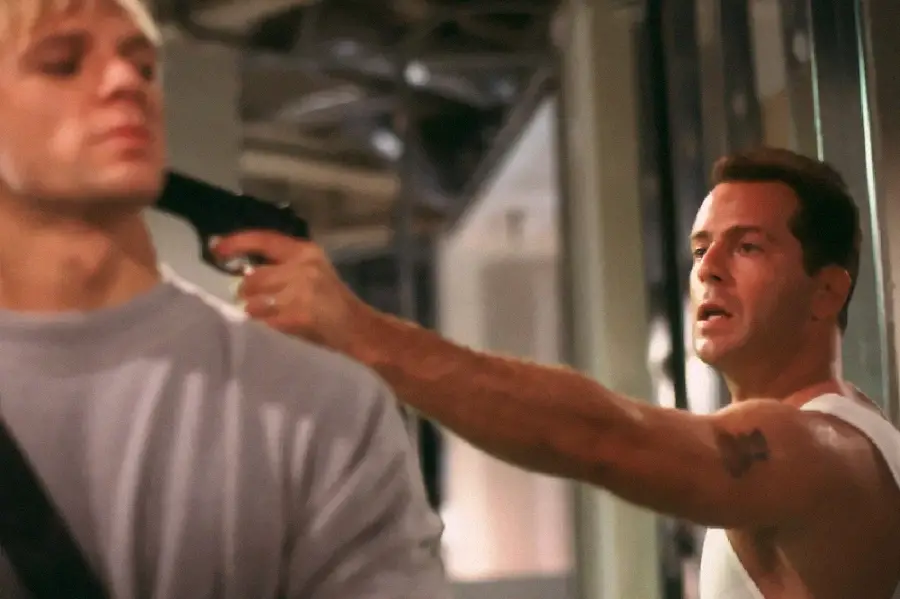 A man in a white tank top holds a gun to the back of another man's head inside an industrial or office setting; both appear tense and focused.