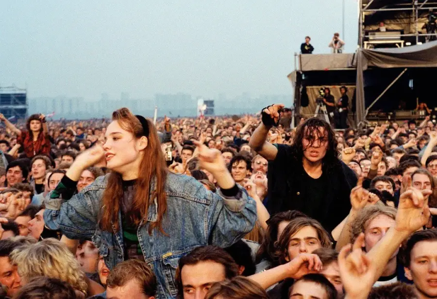 A large crowd of enthusiastic people at an outdoor concert, with a woman in a denim jacket cheering on someone’s shoulders and others raising their hands and enjoying the music. A stage and scaffolding are visible in the background.