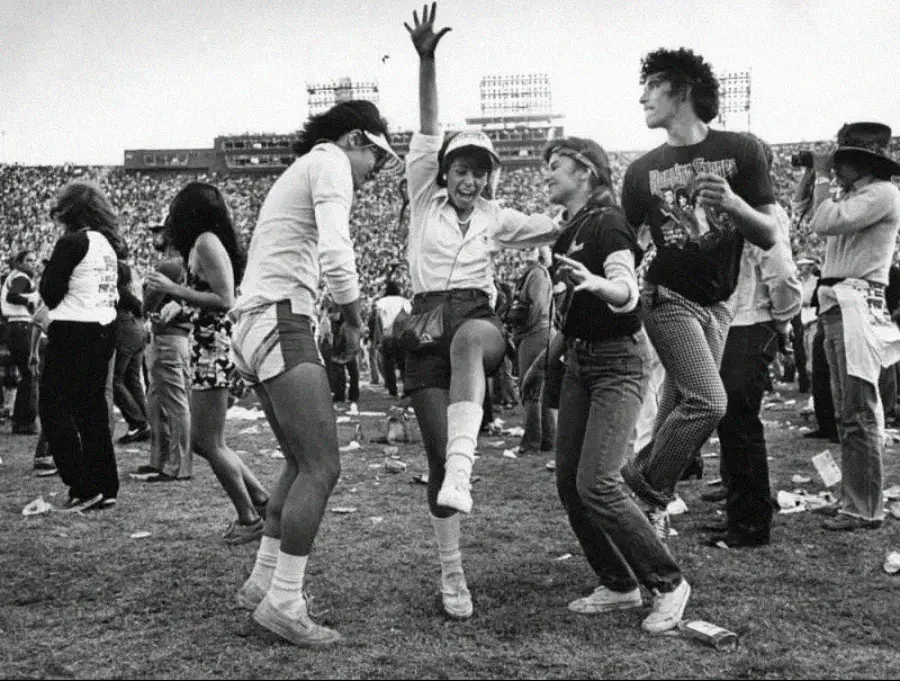 A group of young people dance and celebrate on a grassy field at an outdoor concert or festival, with a large crowd and stadium seats visible in the background.