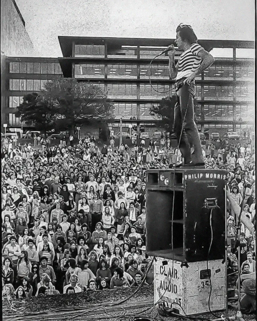 A performer with long hair sings into a microphone while standing on top of speakers labeled “Philip Morris” and “C.A.I.R. Audio,” facing a large outdoor crowd in front of a modern building.